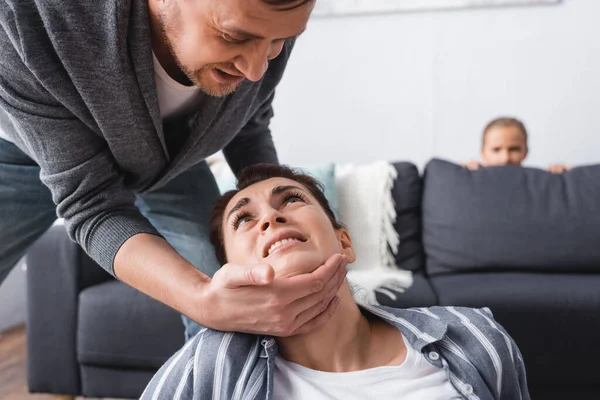 Agresivo marido tocando el cuello de la esposa asustada mientras el niño se esconde detrás del sofá en un fondo borroso - foto de stock