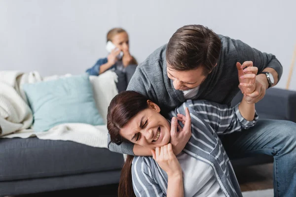 Angry man choking wife while child talking on smartphone on blurred background — Stock Photo