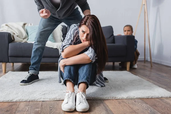 Mujer asustada sentada en el suelo cerca de marido abusivo señalando con el dedo y la hija escondida detrás del sofá sobre fondo borroso — Stock Photo