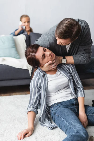 Abusive husband choking wife with closed eyes while kid talking on smartphone on blurred background — Stock Photo