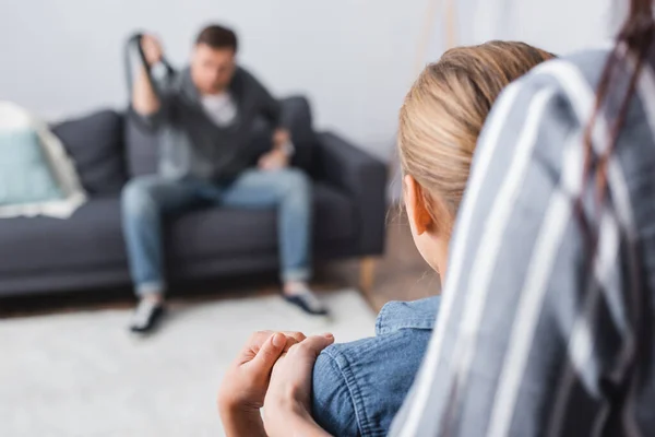 Donna abbracciare bambino mentre abusa marito seduto sul divano su sfondo sfocato — Foto stock