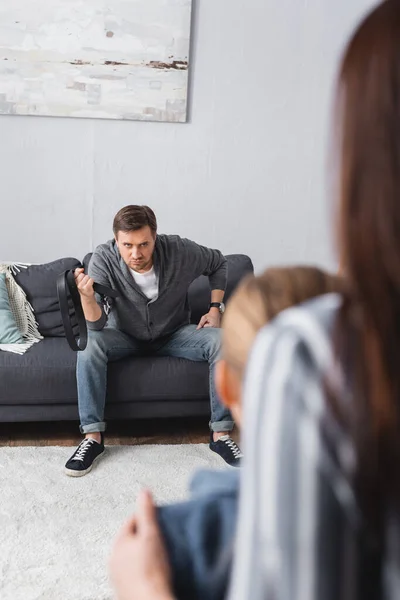 Abusive man holding waist belt near wife and daughter on blurred foreground — Stock Photo