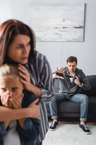 Abusador con cinturón de cintura mirando a esposa asustada abrazando a hija en primer plano borroso — Stock Photo