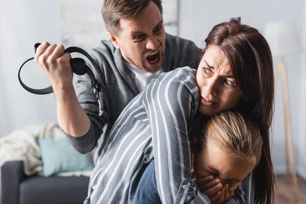 Homme agressif tenant la ceinture de taille et criant à la femme effrayée cachant enfant à la maison — Photo de stock