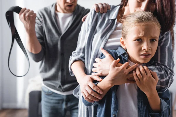 Enfant effrayé embrassant la mère près du père en colère tenant la ceinture sur fond flou — Photo de stock