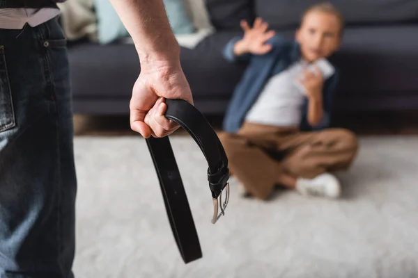 Hombre sosteniendo cinturón de cintura cerca de niño en el suelo en casa sobre fondo borroso - foto de stock