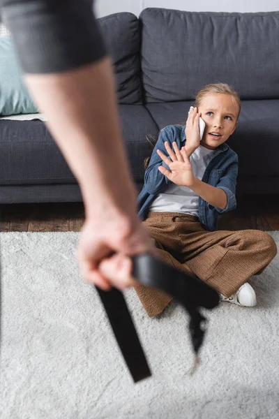 Ängstliches Mädchen spricht auf Smartphone und zeigt Vater Stoppgeste mit Taillengurt auf verschwommenem Vordergrund — Stockfoto