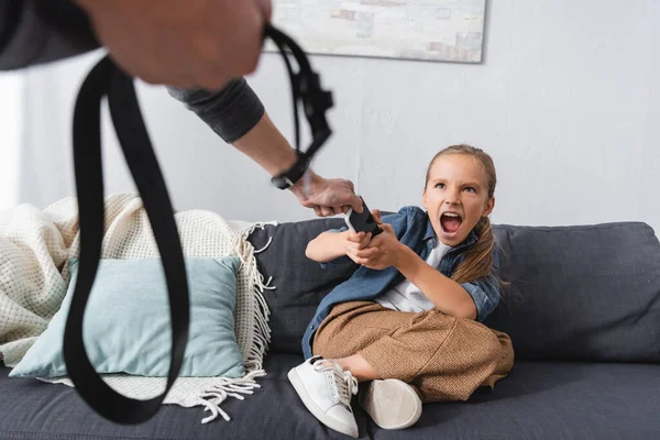 Chico gritando sosteniendo teléfono inteligente cerca del padre con cinturón en primer plano borroso - foto de stock