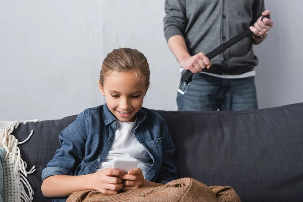Smiling kid using smartphone near father with waist belt on blurred background at home — Stock Photo