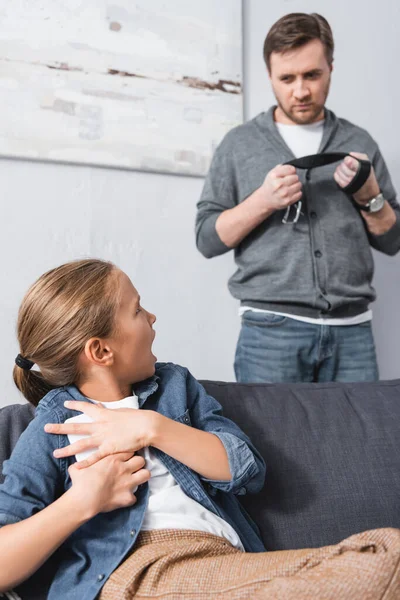 Enfant effrayé cachant smartphone et regardant père avec ceinture sur fond flou — Photo de stock