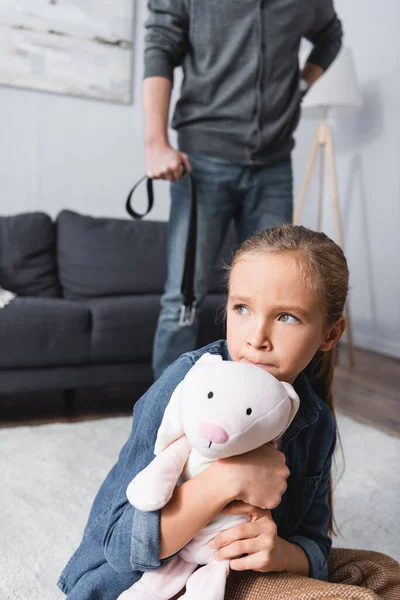 Verängstigtes Mädchen umarmt Stofftier in der Nähe des Vaters mit Taillengurt auf verschwommenem Hintergrund zu Hause — Stockfoto