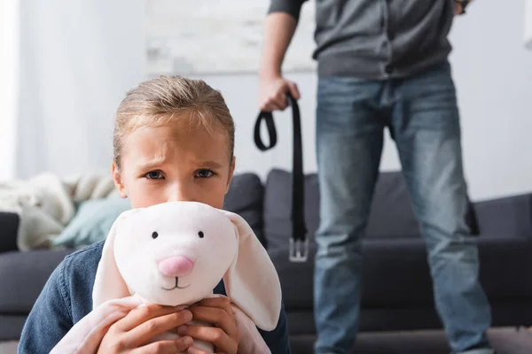 Scared girl with soft toy looking at camera near abusive father with waist belt on blurred background — Stock Photo