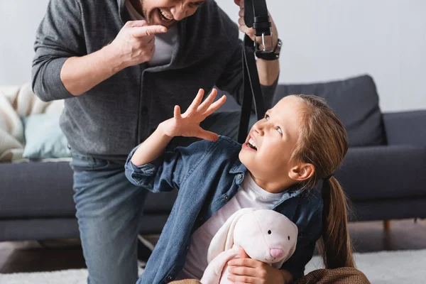 Enfant effrayé avec un jouet doux regardant le père agressif avec une ceinture pointant du doigt sur un fond flou — Photo de stock