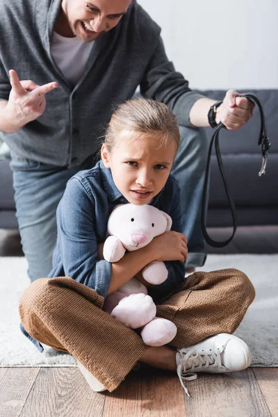 Criança assustada segurando brinquedo macio perto de pai zangado com cinto de cintura no fundo embaçado — Fotografia de Stock