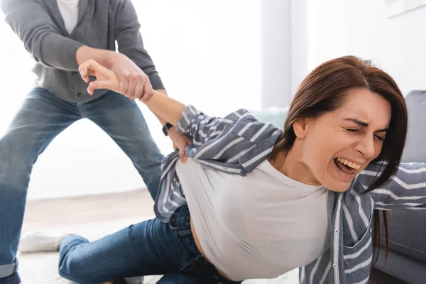 Donna spaventata sdraiata sul pavimento vicino abusatore afferrando le mani su sfondo sfocato a casa — Foto stock