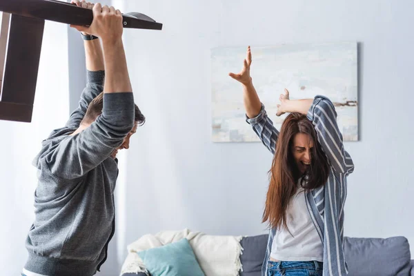 Abusive man holding chair near scared wife covering head at home — Stock Photo