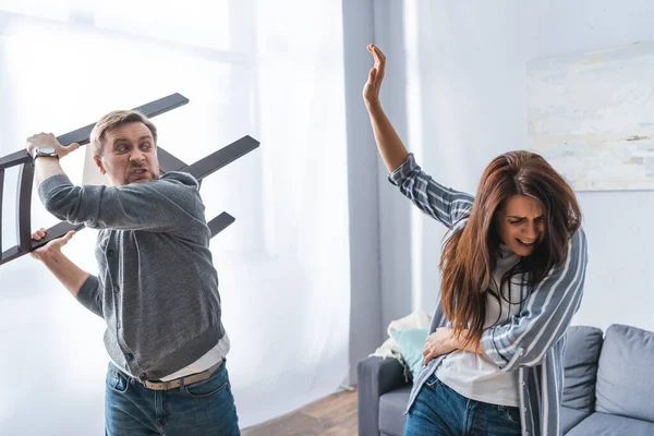 Angry man holding chair near frightened wife at home — Stock Photo