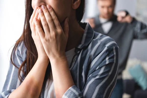 Victim of domestic violence with bruises on hands covering mouth near husband on blurred background — Stock Photo