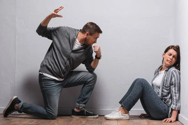 Angry man with hand in fist looking at wife with bruises on floor — Stock Photo