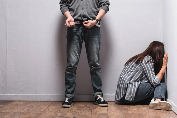 Злоумышленник снимает пояс рядом с женщиной с синяком на руке, сидя на полу возле стен — стоковое фото