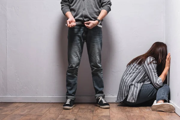 Abuseur décoller ceinture près de la femme avec ecchymose sur la main assis sur le sol à côté des murs — Photo de stock