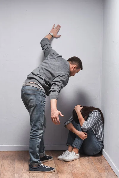 Abusive husband standing near wife with bruises sitting on floor near walls — Stock Photo