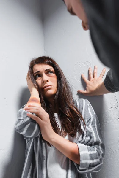 Frightened woman with bruises on face and hand near head looking at husband on blurred foreground — Stock Photo