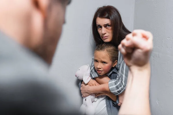 Mujer asustada abrazando a la hija con moretones y juguete suave cerca de las paredes y el marido mostrando el puño en primer plano borroso - foto de stock