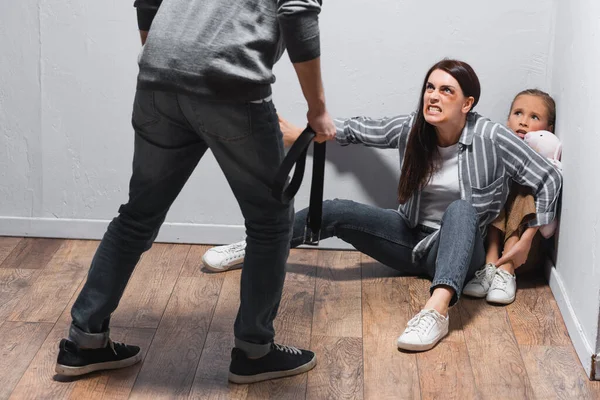 Angry woman with bruises on face hiding child on floor near husband with waist belt on blurred foreground — Stock Photo