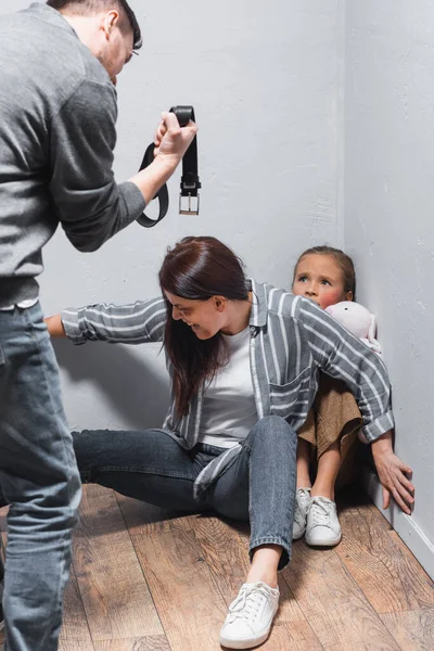 Mujer gritando escondiendo niño cerca de marido con cinturón en primer plano borroso - foto de stock