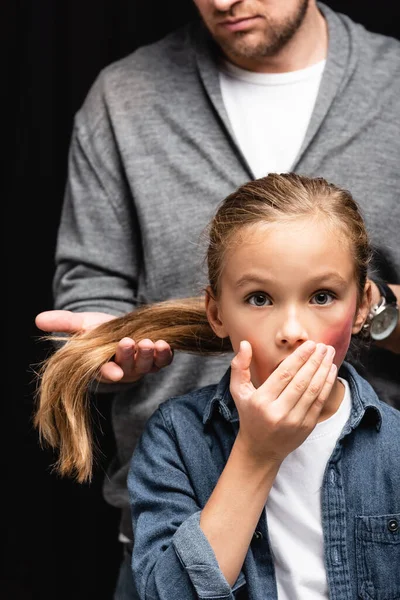 Verängstigtes Kind mit blauen Flecken im Gesicht, das in der Nähe des missbräuchlichen Vaters steht und Haare auf verschwommenem Hintergrund berührt, isoliert auf schwarz — Stockfoto