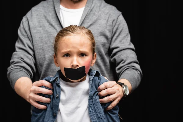 Chica asustada con moretones y cinta adhesiva en la boca de pie cerca de padre sobre fondo borroso aislado en negro - foto de stock