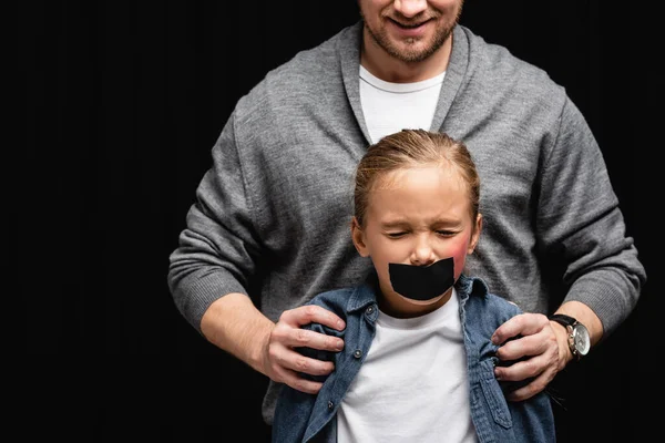 Sonriente abusada abrazando hija con cinta adhesiva y moretones aislados en negro - foto de stock