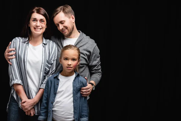 Smiling abused hugging daughter and wife with bruises isolated on black — Stock Photo