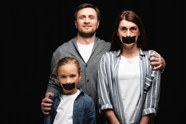 Abusador sonriente abrazando a esposa e hija con moretones y cintas adhesivas aisladas en negro - foto de stock