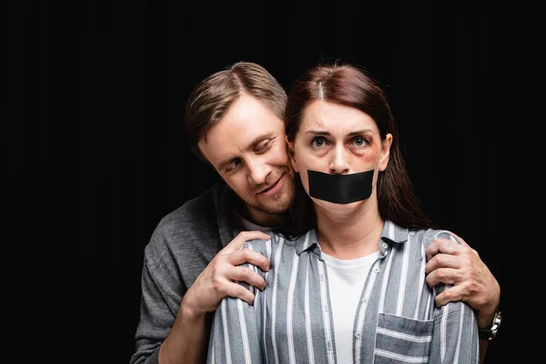 Smiling abuser hugging wife with hematoma and adhesive tape isolated on black — Stock Photo