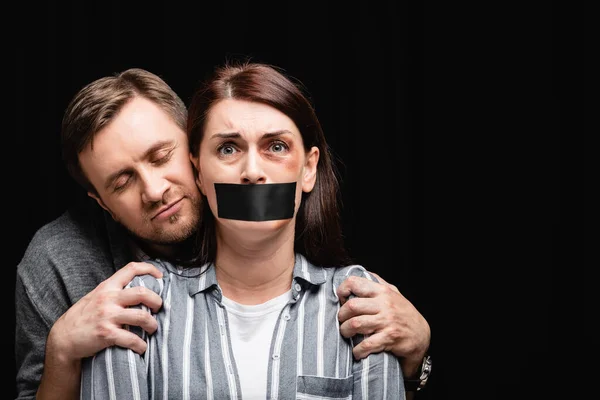 Испуганная женщина с гематомой и клейкой лентой на рту, стоящая рядом с насильником, изолированным на черном — стоковое фото