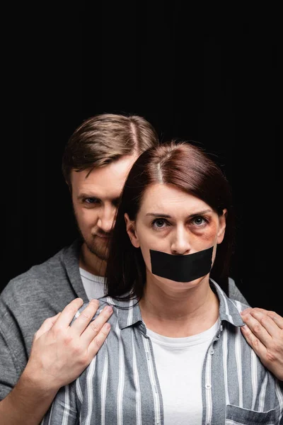 Abuser hiding behind wife with bruise and adhesive tape on mouth isolated on black — Stock Photo