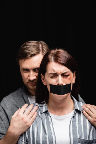 Abusive husband embracing depressed wife with adhesive tape and bruise isolated on black — Stock Photo