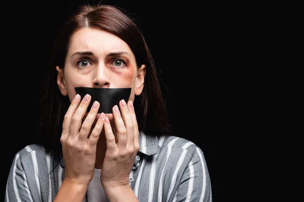 Scared woman in bruises touching adhesive tape on mouth isolated on black — Stock Photo