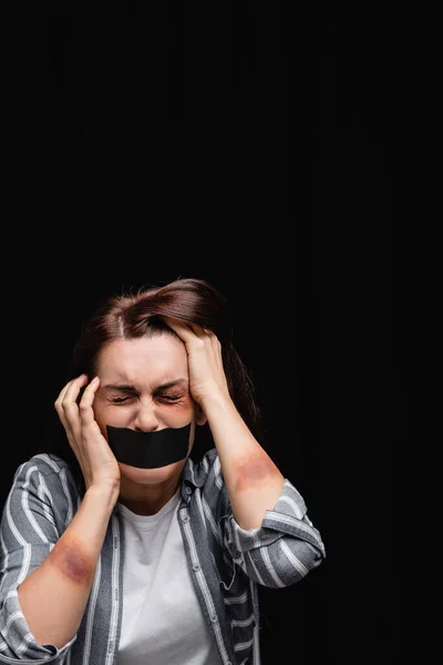 Mulher deprimida com hematomas e fita adesiva na boca isolada em preto — Fotografia de Stock