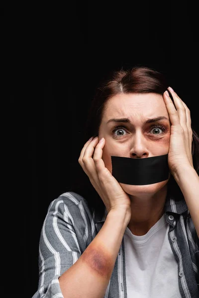 Frightened victim of domestic violence with adhesive tape on mouth looking at camera isolated on black — Stock Photo