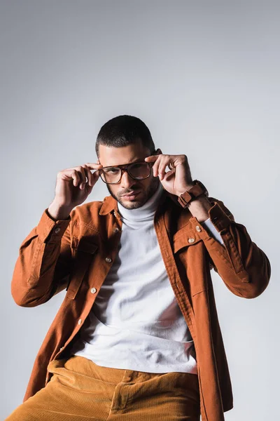 Young man looking at camera while adjusting eyeglasses isolated on grey — Stock Photo