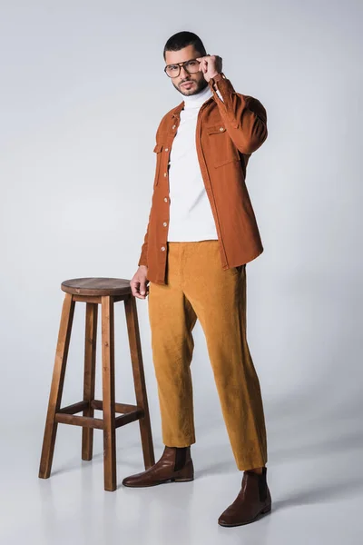 Elegante hombre sosteniendo anteojos cerca de silla de madera sobre fondo gris - foto de stock