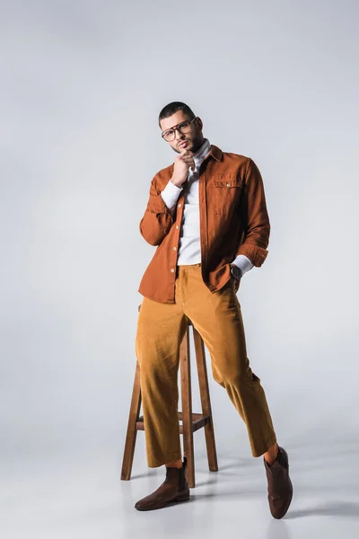 Homme à la mode avec la main dans la poche de pantalon regardant la caméra près de la chaise sur fond gris — Photo de stock