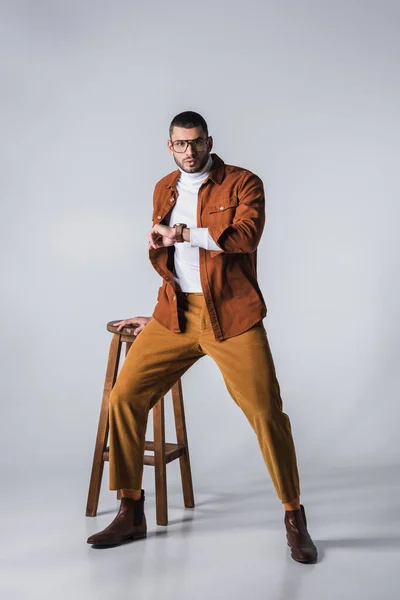 Hombre con estilo en gafas y reloj de pulsera mirando a la cámara cerca de la silla sobre fondo gris - foto de stock