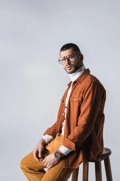 Homme élégant en lunettes et veste en terre cuite assis sur une chaise isolée sur gris — Photo de stock