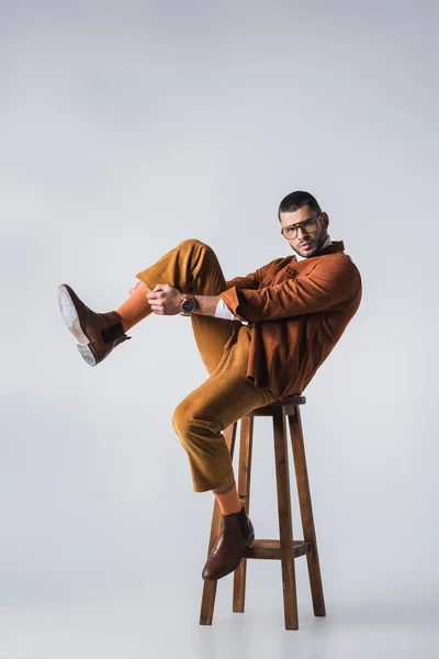 El hombre de moda ajustando calcetín naranja mientras está sentado en la silla sobre fondo gris - foto de stock