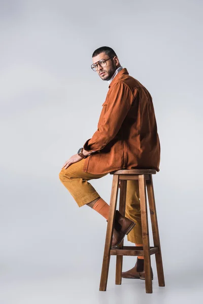 Stylish man in eyeglasses and terracotta jacket looking at camera on wooden chair on grey background — Stock Photo