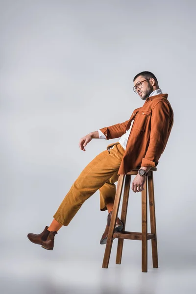 Stylish man in eyeglasses and terracotta jacket posing on wooden chair on grey background — Stock Photo